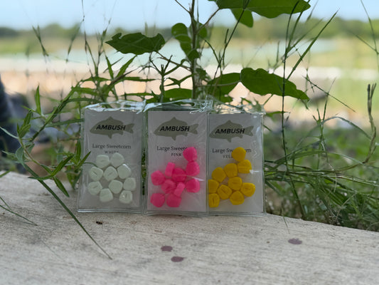 Three small clear plastic packages of Ambush live bait fish, containing white, pink, and yellow bait pieces, displayed outdoors on a stone surface with green grass and foliage in the background.