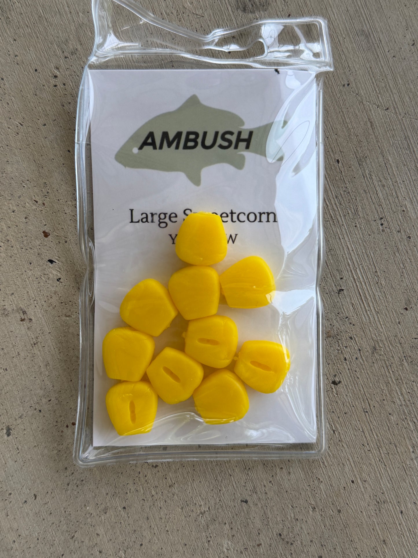 A clear plastic bag containing ten bright yellow corn-shaped fishing lures, attached to a white backing card printed with the brand name 'AMBUSH' and product text, placed on a textured gray surface.
