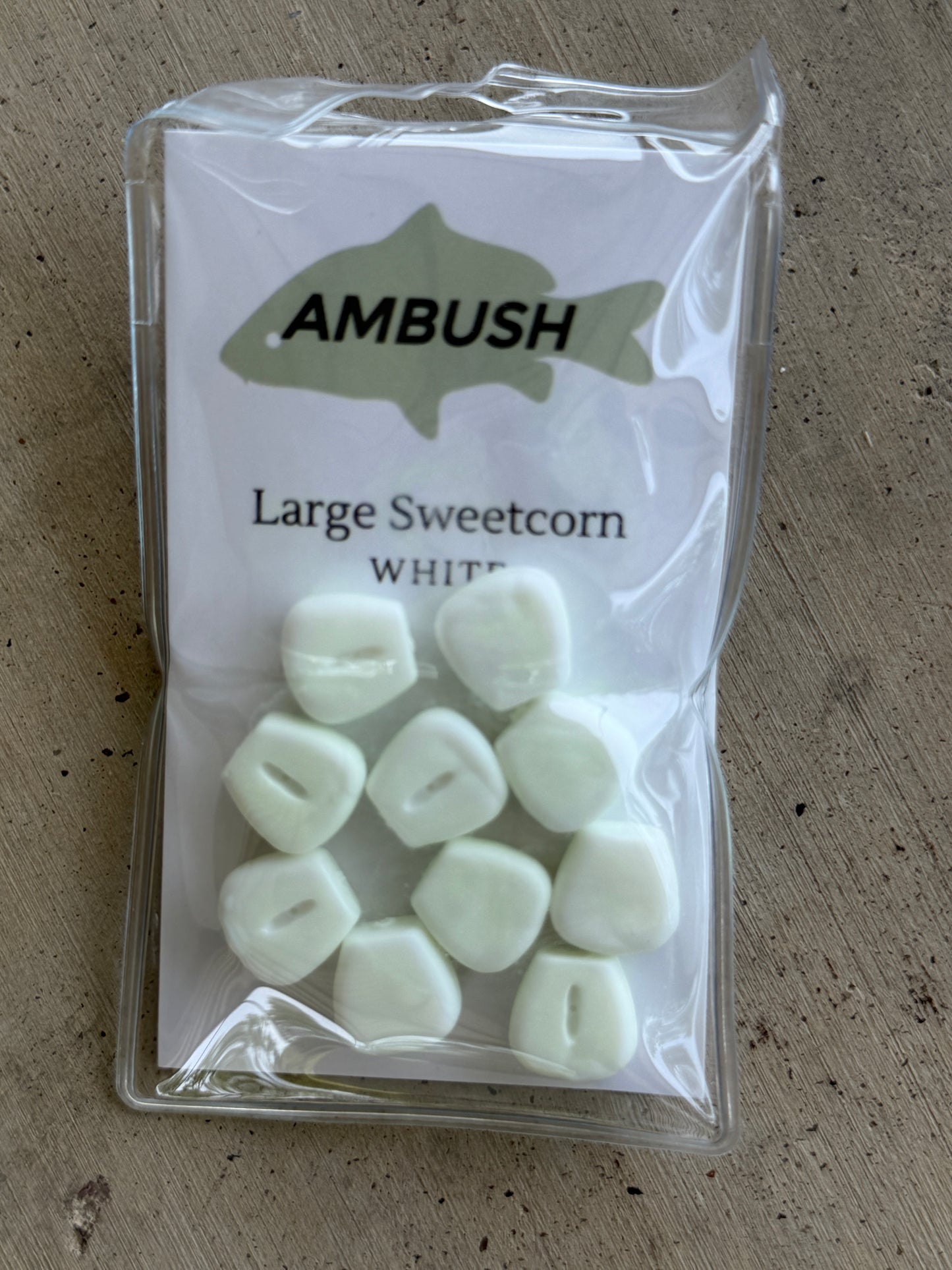 A clear plastic bag containing white sweetcorn cobs, labeled 'AMBUSH Large Sweetcorn WHITE', placed on a textured gray surface.