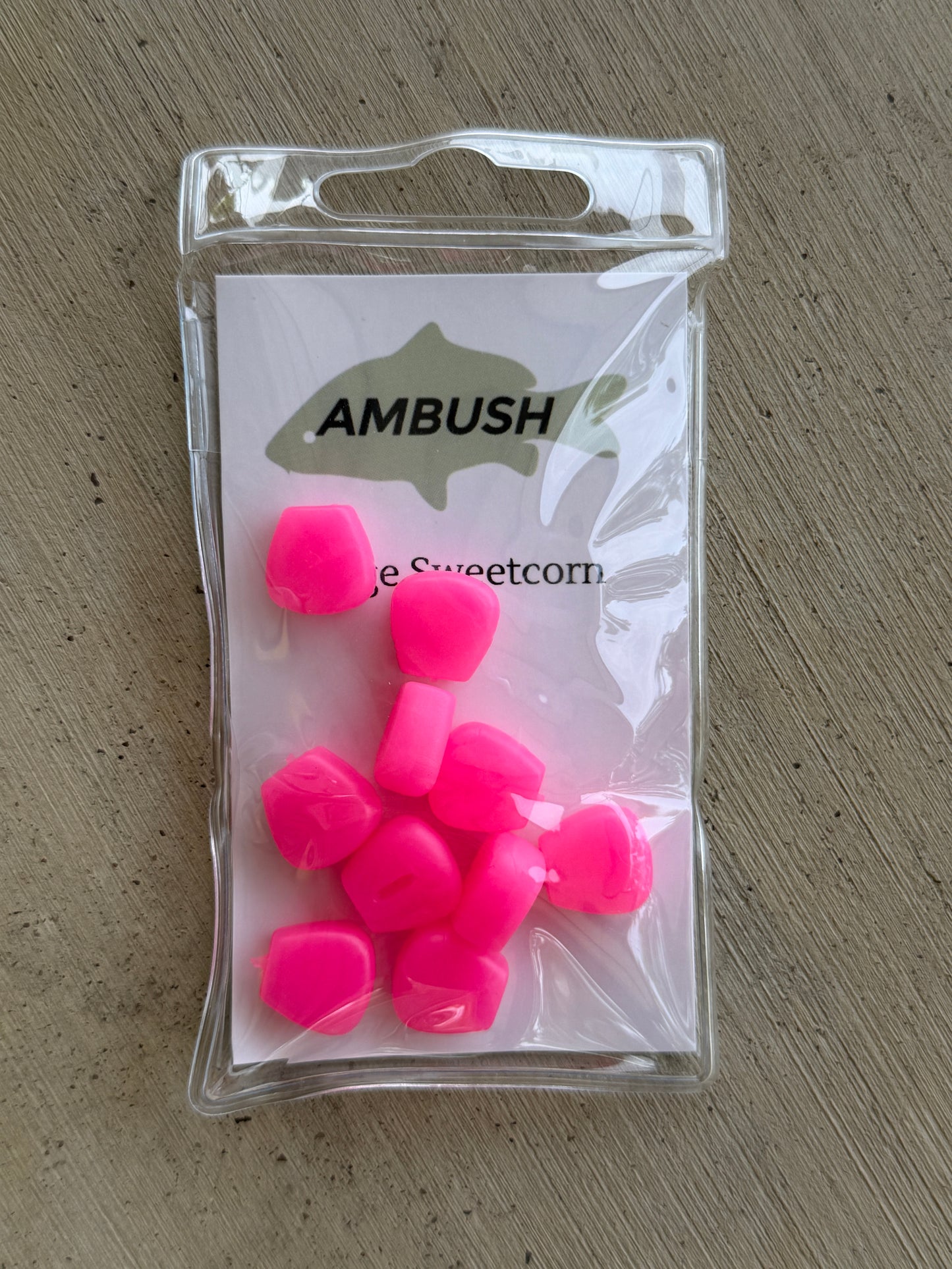 A clear plastic bag containing several bright neon pink fishing lures, with a white backing card printed with the brand name 'AMBUSH' and a fish silhouette, placed on a gray textured surface.