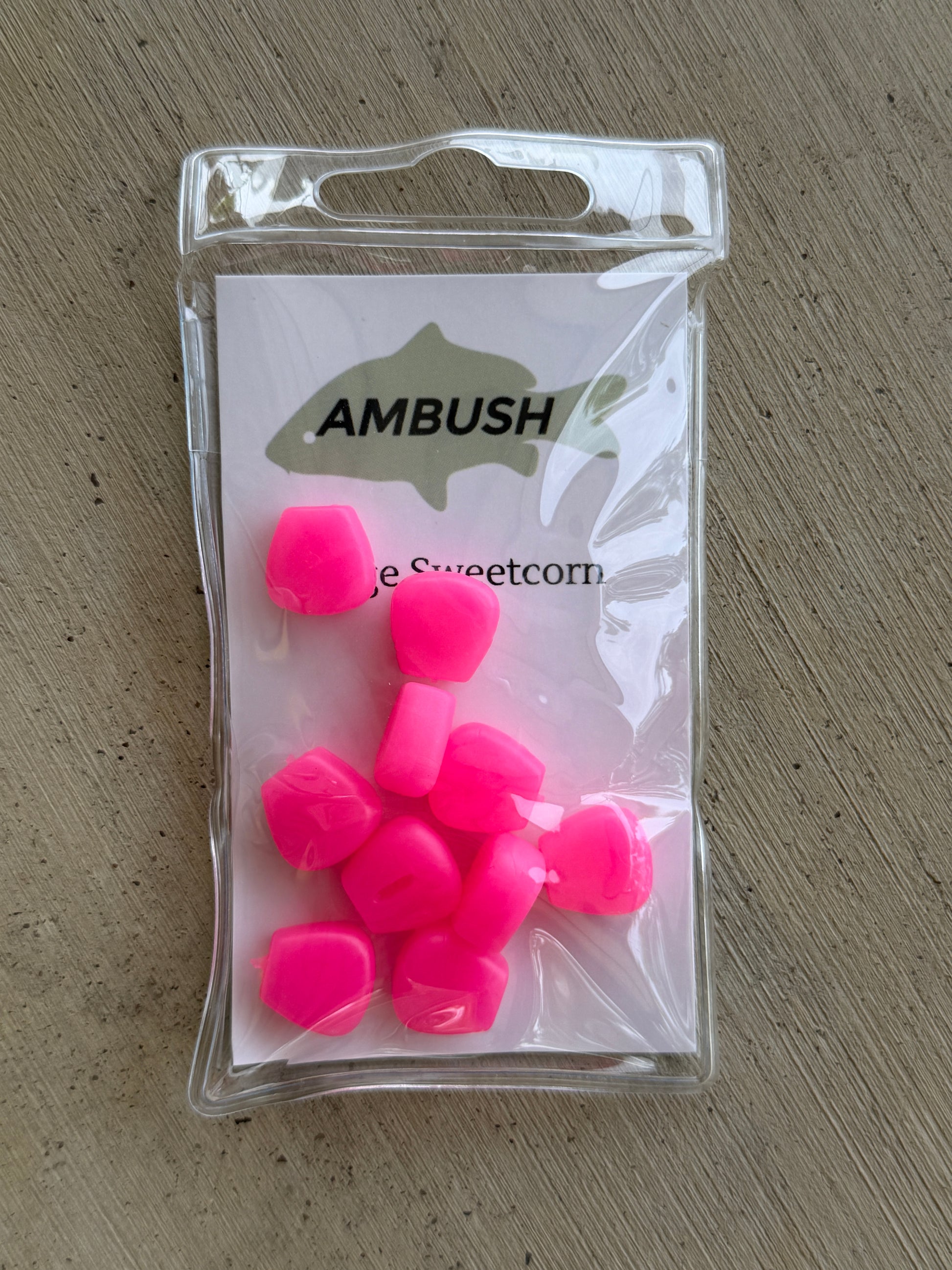 A clear plastic bag containing several bright neon pink fishing lures, with a white backing card printed with the brand name 'AMBUSH' and a fish silhouette, placed on a gray textured surface.